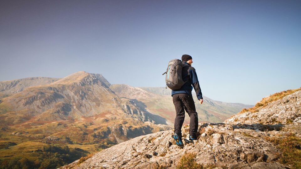 A lone make hiker with backpack walks away from the camera and toward the Welsh mountains.